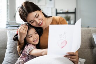 Child daughter is congratulating mom and giving her postcard. Mum and girl smiling and hugging.